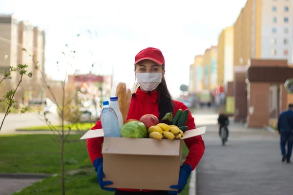 Sokakta bir kutu yiyecek taşıyan kurye kız. Coronovirüs salgını. Koruyucu maskeli ve eldivenli kız içinde sebze, meyve, ekmek, su, süt olan bir kutu tutuyor..