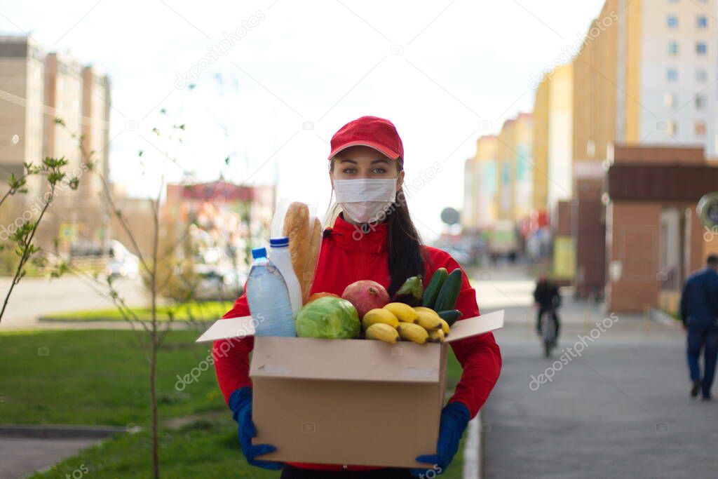 Courier girl with a box of food on the street. Coronovirus pandemic. The girl from the delivery service in a protective mask and gloves holds a box in which are: vegetables, fruits, bread, water, milk.