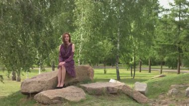 Parktaki bir kızın portresi. Ağır çekim 2x. Leylak elbiseli tatlı kız bir taşın üzerinde oturuyor. Rüzgar esiyor.