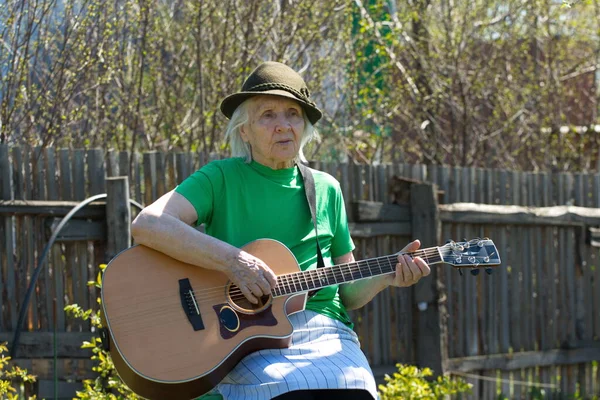 Portrait of a grandmother in nature. Grandmother plays acoustic guitar in the spring garden. She is sitting near an old wooden fence.