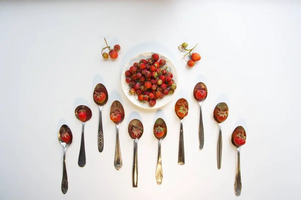 Wild strawberry (alpine strawberry). Top view. Strawberries in teaspoons and in a saucer on a white background. Two branches with berries near a saucer.
