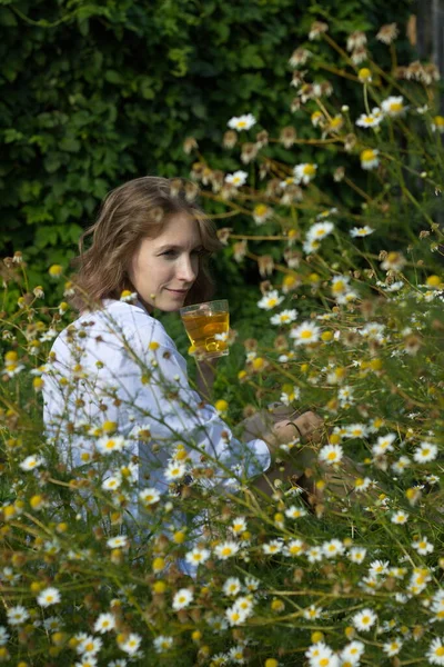 Doğada çay içmek. Genç bir kadın papatya çayının arasında oturuyor ve elinde bir kupa çay tutuyor..