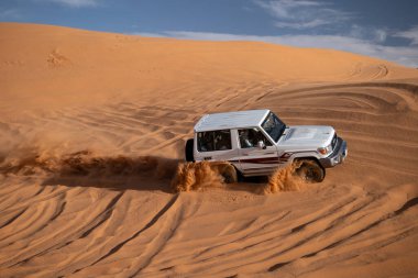 4x4 SUV çöl kumullarında sürüyor, Suudi Arabistan. Beyaz 4x4 SUV arabası Suudi Arabistan 'daki Arap çölünün altın kum tepelerinden geçiyor. Off-road macerası ve mavi gökyüzünün altında çöl keşfi