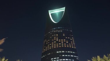 Riyad, Suudi Arabistan 'daki Krallık Merkez Kulesi. Riyad, Suudi Arabistan 'daki palmiye ağaçları tarafından çerçevelenmiş ikonik Krallık Merkezi Kulesi' nin gece görüşü. Aydınlatılmış gökdelen en büyük şehirlerden biridir.