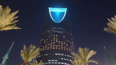 Riyad, Suudi Arabistan 'daki Krallık Merkez Kulesi. Riyad, Suudi Arabistan 'daki palmiye ağaçları tarafından çerçevelenmiş ikonik Krallık Merkezi Kulesi' nin gece görüşü. Aydınlatılmış gökdelen en büyük şehirlerden biridir.