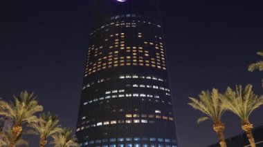 Riyad, Suudi Arabistan 'daki Krallık Merkez Kulesi. Riyad, Suudi Arabistan 'daki palmiye ağaçları tarafından çerçevelenmiş ikonik Krallık Merkezi Kulesi' nin gece görüşü. Aydınlatılmış gökdelen en büyük şehirlerden biridir.