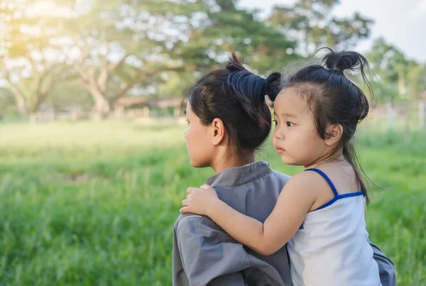 Portrait of an Asian cute little girl hugs aunt\'s shoulders on piggybacks and looks straight to the green meadow during summer holidays with copy space for text. Happiness and love of parenthood, family concept.