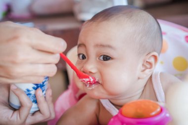 Bebek sandalyesinde bebek maması yiyen neşeli şirin bir bebek. Anne bebeğini kırmızı kaşıkla besliyor. Evde mutlu bir Asyalı aile ve ev hayatı. Çocuklar için sağlıklı beslenme