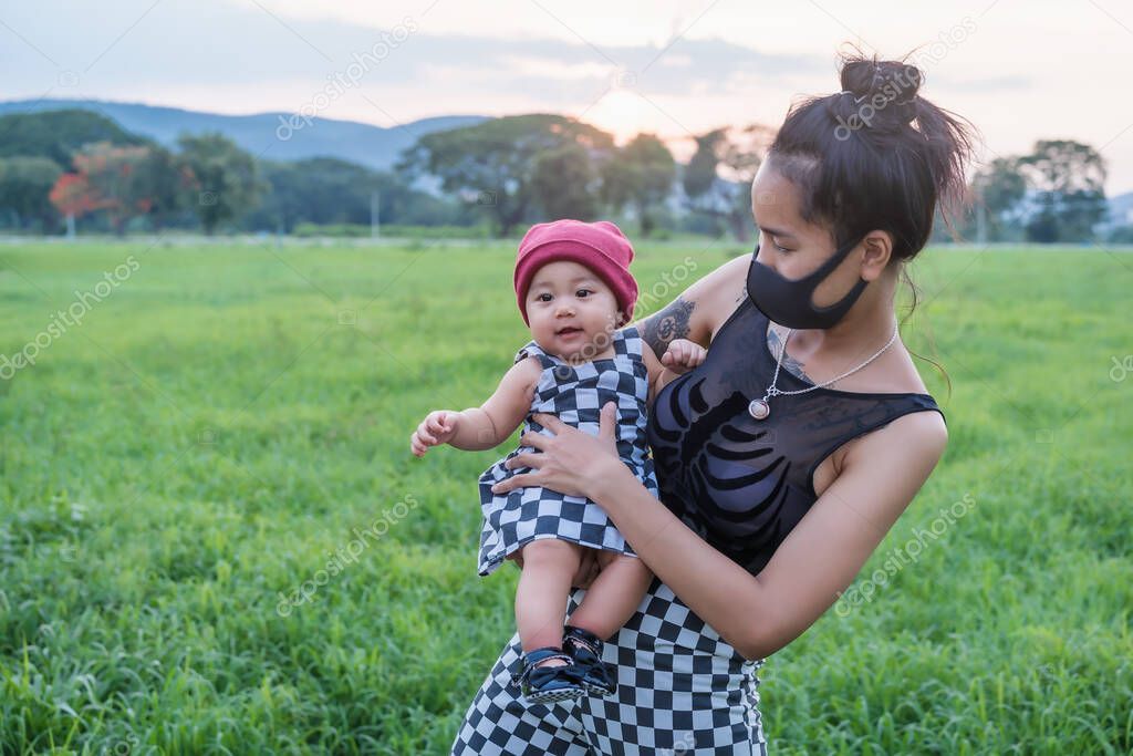Portrait of a happy Asian mother wears black mask and plays with her young daughter in the summer garden at sunset. Healthcare and social distancing activity for the Covid-19 epidemic concept