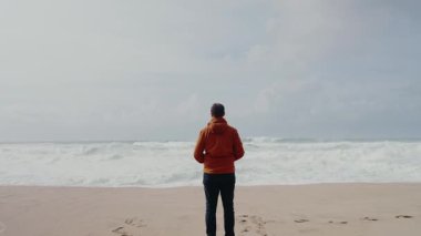 A man in an orange jacket stands on a sandy beach, facing the ocean. The waves crash gently, reflecting a calm and contemplative mood. Man in Orange Jacket Standing Alone on Foggy Beach