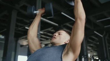 A determined man performs shoulder press exercises with dumbbells in a modern gym setting. Building strength and focusing on fitness goals. Focused Man Lifting Weights In Gym For Strength Training