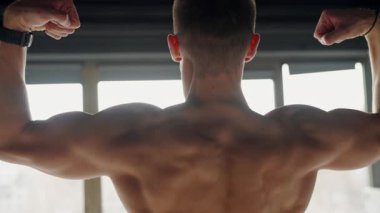 Rear view of a young male athlete using resistance bands to strengthen his back muscles. Focus is on his toned muscles in a bright gym setting. Male Athlete Building Muscle Strength