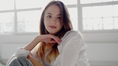 Portrait of a young woman with a subtle smile, comfortably seated in a light-filled room, exuding a relaxed and happy vibe. Young Woman Relaxing in a Bright Cozy Room, Casual and Content