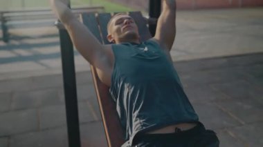 A young man is resting on an outdoor workout bench under the sun. The serene atmosphere captures a moment of relaxation and fitness in a park setting. Young Man Relaxing on Outdoor Workout Bench