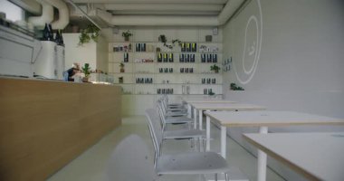 A spacious and well-lit cafeteria featuring a modern design with white chairs, wooden tables, and a barista station. The room is decorated with green plants and shelving filled with various items