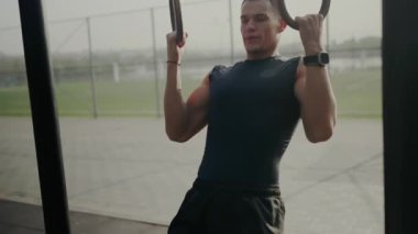 A focused young man exercises using gymnastic rings at an outdoor training facility, displaying strength and endurance. Young Male Athlete Training with Gymnastic Rings Outdoors