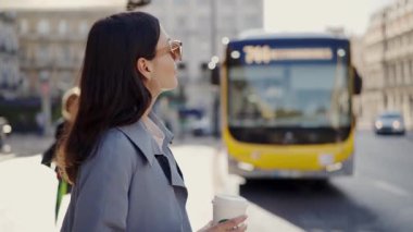 A young adult woman in a trench coat holds a coffee cup, waiting for a bus on a sunny city street, depicting urban life and routine. Young Woman Holding Coffee Waiting for Bus in Urban Setting