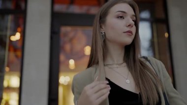 An image capturing an elegant young woman dressed in a stylish beige blazer, confidently posing in an urban, evening setting with soft lights in the background.