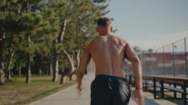 A shirtless man runs energetically outdoors by a colorful soccer stadium, showcasing fitness and vitality in an energetic environment surrounded by nature. Athletic Man Running Outdoor