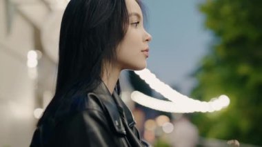 Portrait of a beautiful woman in a leather jacket, standing outdoors during twilight. The image captures a contemplative and serene mood with blurred city lights in the background.