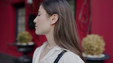 A poised young woman in a white blouse stands confidently in front of a vibrant red door, her expression contemplative and serene. Elegant Young Woman Posing in Front of a Red Door in the City