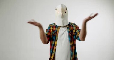 A man wearing a bird mask shrugs his shoulders. He is confused and unsure. He gestures with his hands, palms up. He is wearing a plaid shirt. The background is white.