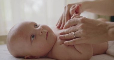 Tombul yanakları ve büyük mavi gözleri olan sevimli yeni doğmuş çocuk vücudunu hareket ettiriyor ve kameraya bakıyor. Güzel, yeni doğmuş bebek yatak çarşaflarında yatıyor, ayak parmaklarına dokunuyor gülüyor kameraya bakıyor..