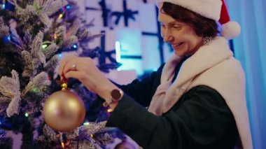 Woman wearing a Santa hat and scarf decorates a Christmas tree with a golden ornament. The festive scene is complemented by colorful lights and a wintry backdrop. Woman Decorating Christmas Tree