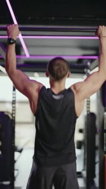 A muscular man performs pull-ups in a modern gym with neon lighting. He focuses on his fitness routine. The gym setting evokes energy and dedication to physical well-being.