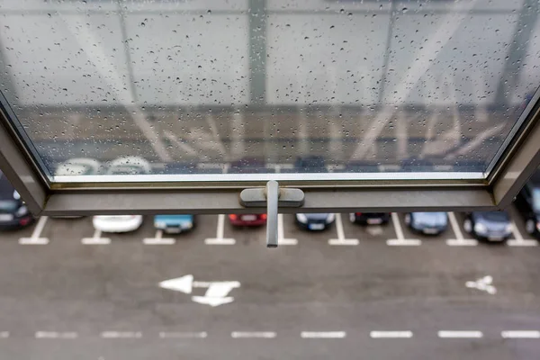 Pencereyi bir sapla aç. Camda yağmur damlaları var. Aşağıda arabaların ve yol işaretlerinin olduğu bir otopark var..