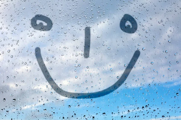 Figure emoticon on the misted glass. Glass window with raindrops against the blue sky. Emitation of children's drawings.