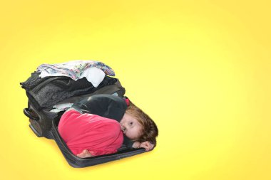 Bir kız eşyaların katlandığı bir bavulun içinde yatıyor. Seyahat kavramı, eşyaların ve kıyafetlerin tasarımı. Sarı arka planda.