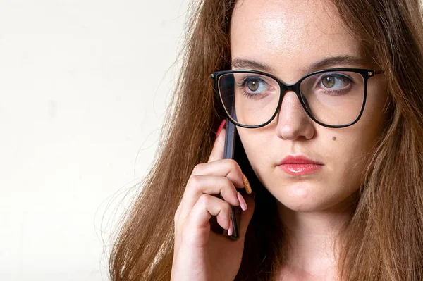 Beautiful girl in glasses is calling on the phone. A pretty girl with long hair brought the phone to her ear and listens. Close-up on a white background, a place for an inscription.