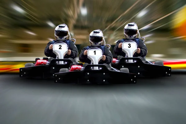 Kapalı alanda kart, hızlı yarışan araba, gokartın olduğu araba, hızlı yarış, yarış afişi. Pist arka planında kart sürüşü yapan üç sürücü var. Boşluğu kopyala.