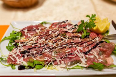 Et carpaccio. İnce dilimlenmiş çiğ et carpaccio, üzerine parmesan peyniri serpiştirilmiş maydanoz ve limon serpiştirilmiş, beyaz tabağa koyu sos serpiştirilmiş. Yakın plan.