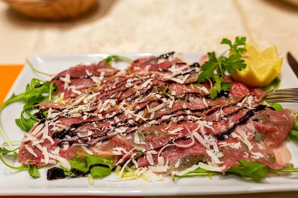 Et carpaccio. İnce dilimlenmiş çiğ et carpaccio, üzerine parmesan peyniri serpiştirilmiş maydanoz ve limon serpiştirilmiş, beyaz tabağa koyu sos serpiştirilmiş. Yakın plan.