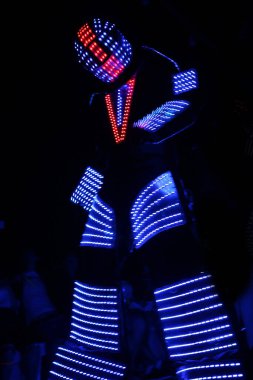 Neon kostüm. Işık çizgileri olan robot kostümlü adam karanlıkta dans ediyor, siyah arka planda parlak takım elbiseli insan siluetleri dans ediyor..
