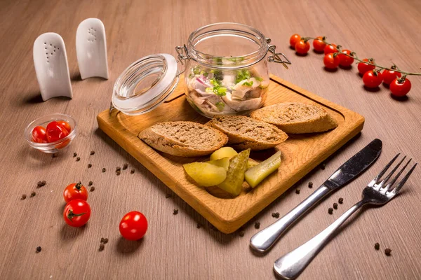 Herring under a pickled onion in a glass jar on a wooden board with slices of bread and pickled cucumber.