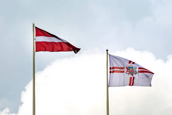 The flag of Latvia and the Presidential flag of Latvia with the coat of arms of Latvia on a long flagpole against the sky. Red-white flag.
