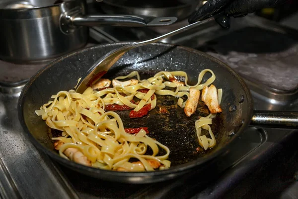 Cooking pasta with red fish and sun-dried tomato. The cook mixes the pasta, which he warms in a black pan.