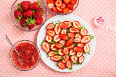 Tabakta tatlı dilimlenmiş çilek, cam kasede çilek reçeli, ve pembe bir benekli masa örtüsünde diğer tatlı meyveler.