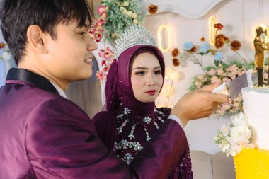 Tören sırasında düğün pastasını kesen görkemli mor elbiseli Endonezyalı Müslüman bir çiftin yakın plan profili. Bu kutlama anı onların yeni başlangıcını temsil ediyor.