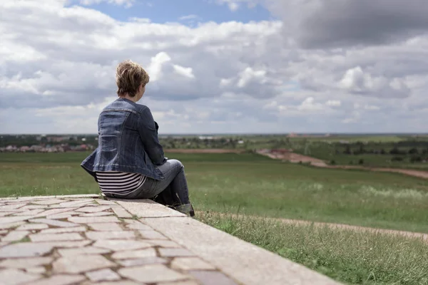 Jeune femme assise sur une tuile de pierre et regardant au loin les champs contre le beau ciel — Photo