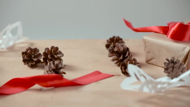 Різдвяні та новорічні концертні подарунки, упаковані в крафт-папір, сніжинки, вирізані з білого паперу, соснові шишки, червоні стрічки на легкому дерев'яному столі . — стокове відео