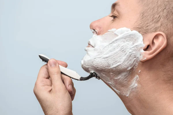 Fotografia de um homem a fazer a barba com uma navalha num fundo cinzento. Vista lateral. Foto com espaço de cópia . — Fotografia de Stock
