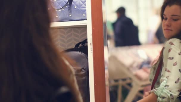 Una mujer joven en una tienda está eligiendo una bolsa, de pie junto al espejo en la tienda — Vídeo de stock