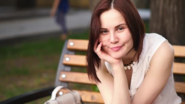 Belle jeune femme sourit mystérieusement, assise sur un banc dans un parc d'été. Portrait d'une femme à l'extérieur . — Video