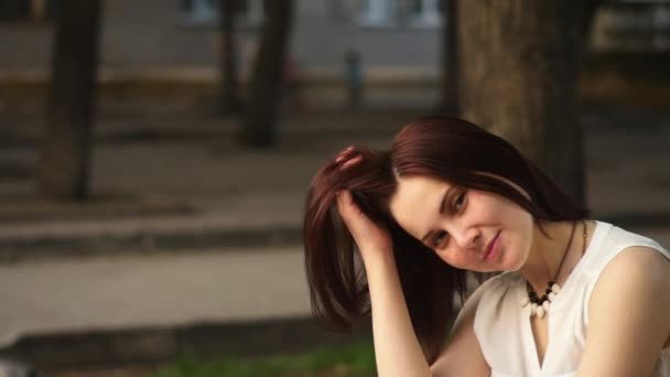 Hermosa joven sonríe misteriosamente, sentada en un banco en un parque de verano. Retrato de una mujer al aire libre . — Vídeos de Stock