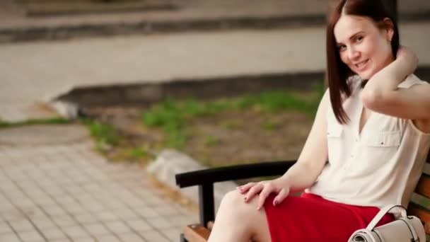 Όμορφη νεαρή γυναίκα χαμόγελα μυστηριωδώς, κάθεται σε ένα παγκάκι στο πάρκο ένα καλοκαίρι. Πορτρέτο μιας γυναίκας σε εξωτερικούς χώρους. — Αρχείο Βίντεο