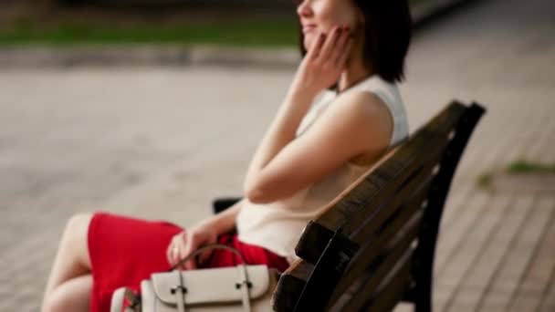 Hermosa joven sonríe misteriosamente, sentada en un banco en un parque de verano. Retrato de una mujer al aire libre . — Vídeos de Stock